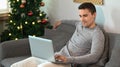 Man sitting on couch near Christmas tree in living room and using laptop computer. Royalty Free Stock Photo