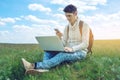 Young man sitting on a green meadow with laptop and using the phone on the background of blue cloudy sky Royalty Free Stock Photo