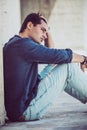Young man sitting on the ground outdoors with hand in hair. Casual clothes, profile view Royalty Free Stock Photo