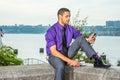 Young man sitting outdoors by river in New York City, holding book, reading cell phone Royalty Free Stock Photo