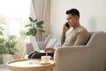 Young man sitting on sofa at home with laptop talking with friend on smartphone Royalty Free Stock Photo