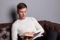 Young man is sitting on a leather sofa and reading a book