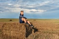 Young guy sitting on a straw stack sadly looking thoughtfully into the distance with a rain sky and a rainbow on the background Royalty Free Stock Photo