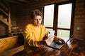 A young man sitting at a table in the country with a smartphone in his hands and uses a laptop in a room with a wooden interior. Royalty Free Stock Photo