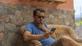 Young man sitting on wicker bench thinking and sending a message. Looking cellphone and planning a solution. Guy with sunglasses Royalty Free Stock Photo