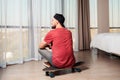 A young man sittingon a longboard, looking out the window. Home interior. Rear view. The concept of quarantine and activity at