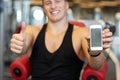 Young man with smartphone showing thumbs up in gym
