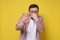 Young man smells an unpleasant smell, he pinches nose with fingers and makes a stop gesture with hand. Yellow background