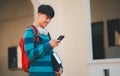 A young man is smiling and looking at his cell phone while holding a backpack