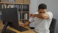 Young man sneezing into his elbow while working at a computer in an office setting filled with file folders and shelves Royalty Free Stock Photo