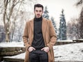 Young man in snowy city park wearing wool coat, looking at camera