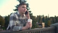 A young man speaks on the phone with a glass of coffee in his hand. A man with a phone near the fence on the embankment