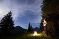 Young man is standing by a burning fire under evening sky with manifesting stars on the background of camping Royalty Free Stock Photo