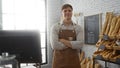 Young man standing confidently in a bakery interior, posing with arms crossed in front of shelves stocked with various breads, Royalty Free Stock Photo