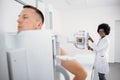 Young man standing in modern hospital during x ray chest scan, while female Afro American medical technician works with