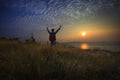 Young man standing and rising hand as victory on grass hill looking to sun above sea horizontal with dramatic colorful sky Royalty Free Stock Photo