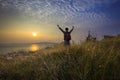 Young man standing and rising hand as victory on grass hill looking to sun above sea horizontal with dramatic colorful sky Royalty Free Stock Photo