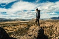 Young man standing on a rock at the top of a cliff overlooking a valley with a mountain range in the background. Concept of Royalty Free Stock Photo