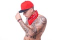 A young man is standing topless in a red cap and a red scarf around his neck with tattoos all over his body