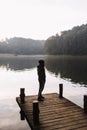 A young man stands alone on a wooden bridge by the lake. Early cloudy foggy morning. To be alone with yourself