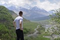 The young man stands high on the mountain next to a flowering tree and looks at the valley and snow-capped mountains