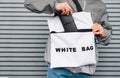 Young man stands outside in stylish clothes against a gray wall background and puts a wallet in a trendy white eco bag.Guy wears a