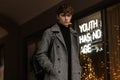 Young man in a stylish gray check coat with a leather trendy backpack stands in the city on the background of a shop window with