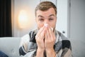 Young man suffering from a common cold and flu or allergy sit at home wrapped in blanket and wipe his nose with tissues Royalty Free Stock Photo