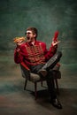 Young man in suit as royal hussar tasting big hot-dog isolated on dark green background. Retro style, comparison of eras