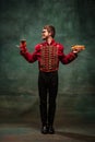 Young man in suit as royal hussar tasting big hot-dog isolated on dark green background. Retro style, comparison of eras
