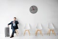 Young man in suit sit on white chair in room alone. He hold hand on suitcase and look at watches. He wait for flight. Royalty Free Stock Photo