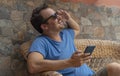 Young man with sunglasses relaxing in wicker chair at terrace. Guy with blue shirt looking up holding his cellphone, sitting and Royalty Free Stock Photo