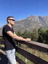 Young man in sunglasses stands on observation deck and looks away in sunny day. Male tourist enjoys beautiful view of