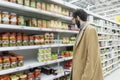 Young man in the supermarket. Shelves with a large selection of canned food. Brunette with a beard in a medical mask in a beige