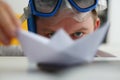 Young man in swimming goggles plays with paper ship at workplace