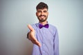 Young man with tattoo wearing purple shirt and bow tie over isolated white background smiling friendly offering handshake as