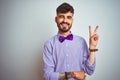 Young man with tattoo wearing purple shirt and bow tie over isolated white background smiling with happy face winking at the Royalty Free Stock Photo