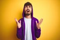 Young man with tattoo wearing purple sweatshirt with hood over isolated yellow background crazy and mad shouting and yelling with