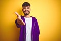 Young man with tattoo wearing sport purple sweatshirt over isolated yellow background smiling friendly offering handshake as