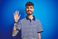 Young man with tattoo wearing striped polo standing over isolated blue background showing and pointing up with fingers number five