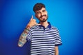 Young man with tattoo wearing striped polo standing over isolated blue background smiling doing phone gesture with hand and Royalty Free Stock Photo