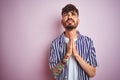 Young man with tattoo wearing striped shirt standing over isolated pink background begging and praying with hands together with