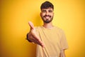 Young man with tattoo wearing striped t-shirt standing over isolated yellow background smiling friendly offering handshake as