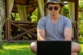 A young man, a teenager, a student, sits in a hat and sunglasses on the green grass, works on a laptop, against the background of