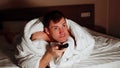 Young man under blanket lying on bed and watching tv in evening. Relaxed male watching programs of show and switches