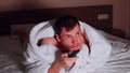 Young man under blanket lying on bed, watching tv and waving his legs in late evening. Relaxed male watching programs of