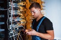 Young man in uniform with measuring device works with internet equipment and wires in server room