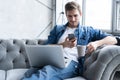Young man using his smartphone for online banking - sitting on sofa with laptop on leap. Royalty Free Stock Photo