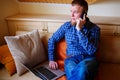 Young man using his smartphone for online banking - sitting on sofa with laptop on leap Royalty Free Stock Photo