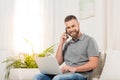 Young man using laptop and talking on smartphone while sitting on sofa and smiling at camera Royalty Free Stock Photo